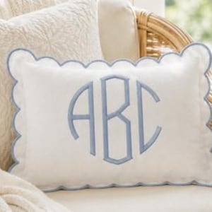 May include: A rectangular white throw pillow featuring a scalloped edge trimmed in light blue piping. The center of the pillow displays a large, embroidered monogram with the letters ABC in a light blue thread, matching the scalloped border.