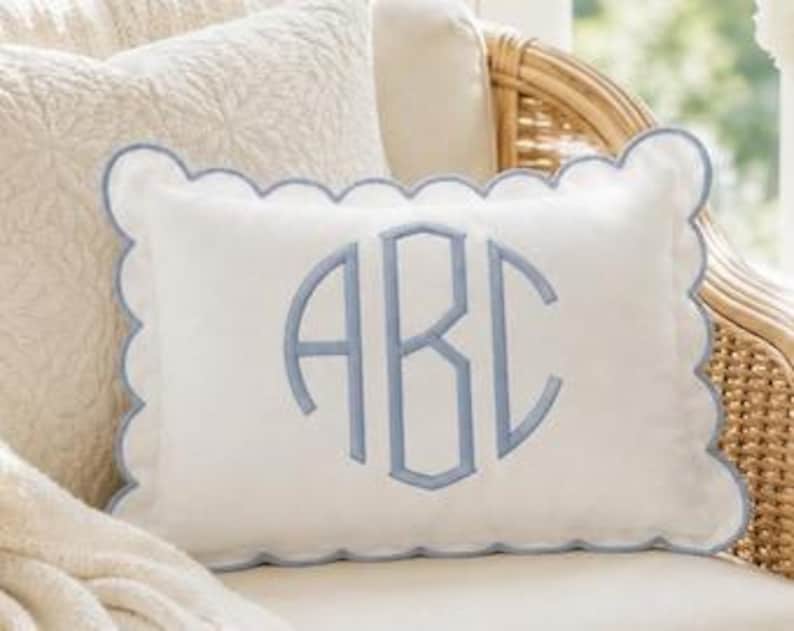 May include: A rectangular white throw pillow featuring a scalloped edge trimmed in light blue piping. The center of the pillow displays a large, embroidered monogram with the letters ABC in a light blue thread, matching the scalloped border.