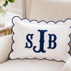 May include: A rectangular white throw pillow featuring a navy blue scalloped edge and a large navy blue embroidered monogram with the letters S, J, and B centered on the front. The pillow has a smooth fabric finish and a decorative border.