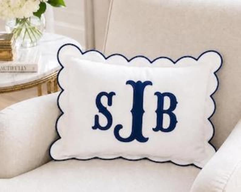 May include: A rectangular white throw pillow featuring a navy blue scalloped edge and a large navy blue embroidered monogram with the letters S, J, and B centered on the front. The pillow has a smooth fabric finish and a decorative border.