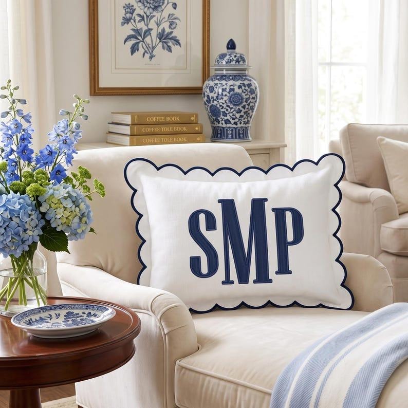 May include: A rectangular white throw pillow with a scalloped edge trimmed in navy blue. The center features a large, embroidered navy blue monogram with the letters SMP in a serif font. The pillow is positioned on a cream-colored armchair.