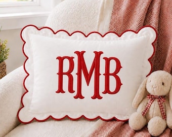May include: A rectangular white throw pillow featuring a scalloped edge trimmed in red piping. The center of the pillow displays the embroidered red monogram letters RMB in a serif font. A small beige plush rabbit toy sits nearby.