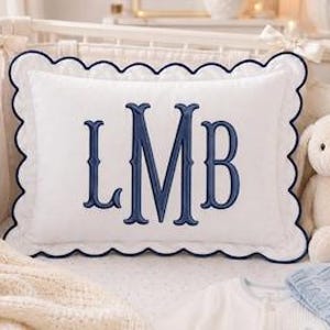 May include: A rectangular white throw pillow featuring a scalloped edge with dark blue piping. The center of the pillow is embroidered with the large dark blue monogram letters LMB in a classic serif font, centered on the white fabric surface.