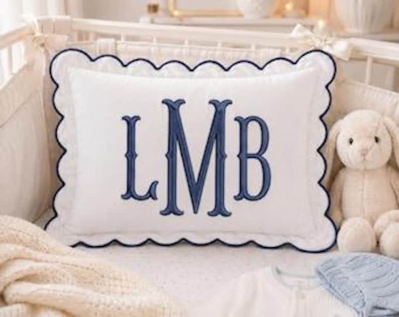 May include: A rectangular white throw pillow featuring a scalloped edge with dark blue piping. The center of the pillow is embroidered with the large dark blue monogram letters LMB in a classic serif font, centered on the white fabric surface.