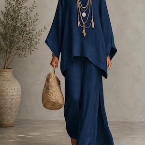 May include: A dark blue two-piece set consisting of a loose-fitting, long-sleeved tunic top with a wide neckline and matching wide-leg trousers featuring side slits. The outfit is accessorised with a multi-strand beaded necklace and a woven straw bag.