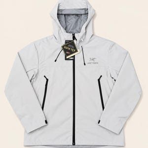 Puede incluir: Una chaqueta con capucha de color gris claro que presenta una cremallera frontal negra de longitud completa, dos bolsillos laterales con cremallera negra y mangas largas con puños ajustables. El logotipo de Arc'teryx está impreso en el pecho y una etiqueta de Gore-Tex cuelga del cuello.