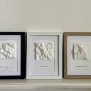 Op de afbeelding: Drie ingelijste wanddecoraties met witte 3D-topografische bergreliëfs. De linkerlijst is zwart, de middelste wit en de rechter is van licht hout. Elk toont de naam van de berg, de hoogte en de locatie in zwarte tekst onder het reliëf.