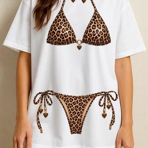 Peut inclure: Un t-shirt blanc à manches courtes avec une impression graphique représentant un bikini à motif léopard. Le haut du bikini comporte une breloque en forme de cœur au centre, et le bas présente des liens latéraux avec des breloques en cœur.