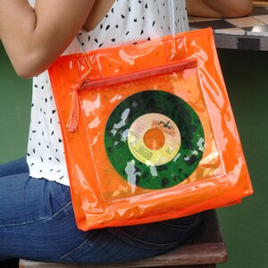Könnte beinhalten: Eine leuchtend orangefarbene, transparente Vinyl-Tragetasche mit Reißverschluss und Quaste. Die Tasche hat ein transparentes Vinyl-Fenster, das eine Vintage-Schallplatte mit einem grün-gelben Etikett zeigt. Das Etikett lautet "Twilight Time" und "The Platters".