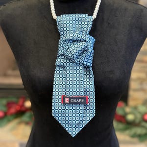 Puede incluir: Una corbata azul claro con un patrón geométrico repetitivo de cuadrados en tonos azul, blanco y negro. La corbata está anudada y muestra una etiqueta rectangular roja y azul marino con el texto blanco CHAPS y un logotipo al frente.