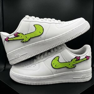 Puede incluir: Un par de zapatillas blancas de corte bajo con un parche bordado personalizado en el lateral. El parche tiene forma de cactus verde lima formando un diseño de swoosh con pequeñas flores rosas en los extremos y detalles de espinas negras.
