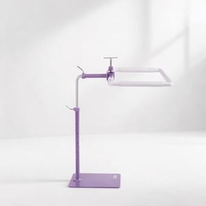 May include: A purple metal embroidery stand with a square base and an adjustable vertical pole. The stand features a horizontal arm attached to a white rectangular embroidery frame, secured by a metal screw mechanism for height and angle adjustment.