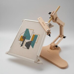 May include: A light-colored wooden embroidery stand with an adjustable arm and a rectangular frame holding a piece of cream-colored fabric. The fabric features a geometric cross-stitch pattern in teal, mustard yellow, and dark gray. The brand name Nurge is visible on the wooden frame and base.