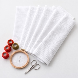 May include: A set of five folded white waffle-weave towels, a round wooden embroidery hoop containing white fabric with two needles, a pair of silver metal scissors, and four small spools of thread in red, orange, and olive green colors.