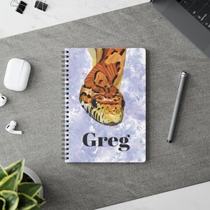 May include: A spiral-bound notebook featuring a graphic illustration of an orange and brown snake on a light purple watercolour background. The name Greg is printed in bold black serif font below the snake illustration on the front cover.