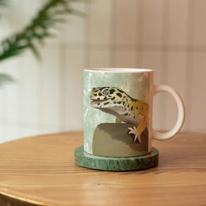 May include: A white ceramic mug featuring a printed illustration of a yellow and black spotted leopard gecko perched on a brown rock against a light green marbled background. The mug has a curved handle and sits on a round green stone coaster.