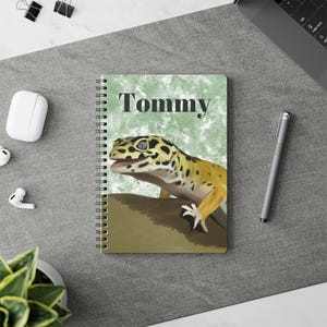 May include: A spiral-bound notebook featuring a yellow and black spotted leopard gecko illustration on the cover. The name Tommy is printed in black text above the gecko, which is set against a textured, light green background.