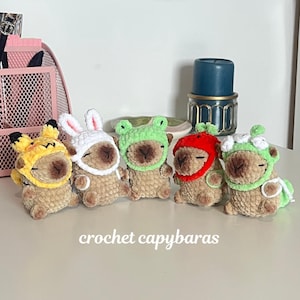 May include: Five small, tan, plush crochet capybaras arranged in a row. Each capybara wears a different crocheted hat: yellow with black-tipped ears, white with long bunny ears, light green, red, and light green with white flower accents.