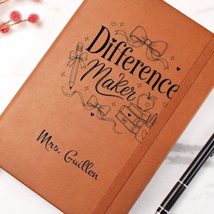 May include: A tan notebook with an elastic closure band and a black pen. The cover features the text "Difference Maker" and "Mrs. Guillen" alongside line art illustrations of a pencil, a bow, an apple, and a stack of books with small hearts.