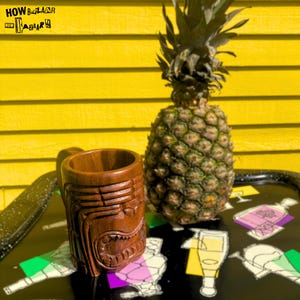 May include: A brown wooden mug carved with a stylized face, positioned next to a whole pineapple on a black tray featuring white line drawings of bottles and glasses. The text "HOW BAZAAR" is visible in the upper left corner of the image.