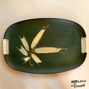 May include: An oval-shaped dark green serving tray with a textured surface. The center features a stylized gold and white leaf or floral graphic. The tray includes two integrated handles wrapped in light-colored, natural-looking woven material.