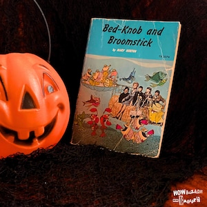 May include: A bright orange plastic jack-o'-lantern with a carved smiling face next to a paperback book titled Bed-Knob and Broomstick by Mary Norton, featuring a light blue cover with an illustration of children and various colorful sea creatures.