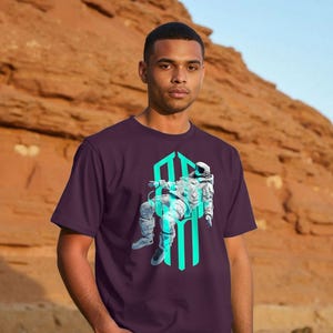 Könnte beinhalten: Ein dunkelviolettes Kurzarm-T-Shirt mit einem Grafikdruck eines im Weltraum schwebenden Astronauten. Der Astronaut ist in Weiß- und Hellblautönen gehalten und befindet sich vor einer vertikalen, leuchtend cyanfarbenen geometrischen Form.