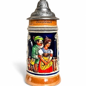 May include: A ceramic beer stein with a metallic lid featuring an embossed floral pattern. The body is tan and dark blue, displaying a relief of two figures in colourful clothing near a fence, with the word Innsbruck printed in black script.
