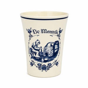 May include: A cream-coloured ceramic cup featuring a dark blue illustration of a person in a hooded robe working near a stone oven. The text "De Monnik" is printed above the scene, which is framed by decorative floral patterns and a thin blue rim.