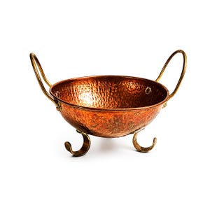 Op de afbeelding: Een ronde kom van gehamerd koper met een gepolijste metalen afwerking, twee gebogen handgrepen aan de zijkanten en drie decoratieve, gekrulde metalen voetjes. De binnen- en buitenoppervlakken hebben een textuurpatroon door het hameren.