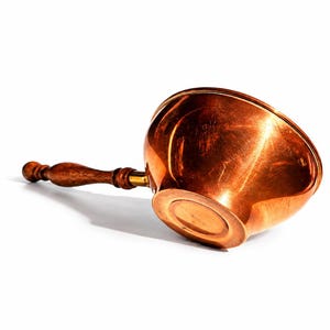 Op de afbeelding: Een gepolijste koperen kom met een afgeronde bodem en een lange, slanke houten steel. De kom heeft een warme, metaalachtige oranjebruine afwerking met zichtbare reflecties. De steel is van donkerbruin, gedraaid hout met een gladde vorm.