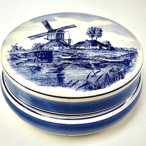 Può includere: Una scatola rotonda in ceramica con coperchio caratterizzata da un design blu e bianco. Il coperchio raffigura un paesaggio con un mulino a vento, una piccola casa e uno specchio d'acqua. La base è blu tinta unita con un bordo bianco superiore.