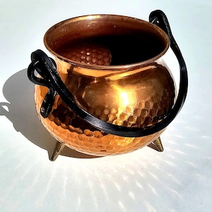 Puede incluir: Un caldero de cobre martillado de forma redonda con una abertura ancha y un interior liso. El recipiente se apoya sobre tres pequeñas patas puntiagudas de color latón e incluye un asa de metal negro gruesa y curva en los laterales.
