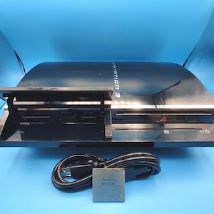 Puede incluir: Una consola de videojuegos PlayStation 3 negra con acabado brillante, que presenta una tapa de ranura para tarjetas multimedia abierta, una ranura para discos y botones de encendido y expulsión. Incluye un cable de alimentación y un chip RSX.