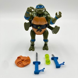 Könnte beinhalten: Eine grüne Schildkröten-Actionfigur mit blauer Maske, orangefarbenem Brustpanzer und schwarzem Gürtel mit L-Emblem. Das Zubehör umfasst eine orangefarbene Wurfscheibe, zwei blaugriffige Schwerter mit silbernen Klingen und einen neongrünen Zylinder.