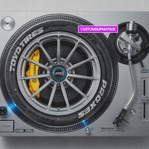 Può includere: Un giradischi Technics argentato con un tappetino personalizzato che presenta la grafica dettagliata di uno pneumatico Toyo Tires Proxes, un cerchio argentato a più razze e una pinza freno Brembo giallo brillante. Il testo CUSTOMSLIPMATSUK è stampato in un riquadro rosa.