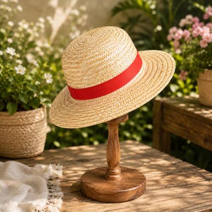 May include: A light tan straw hat with a flat crown and a wide brim, featuring a vibrant red ribbon band wrapped around the base of the crown. The hat is displayed on a dark brown wooden stand with a circular base and a turned pedestal column.