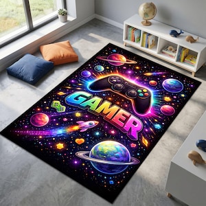 May include: A rectangular area rug featuring a vibrant space-themed design with a black background, colorful planets, stars, a rocket, and a central black game controller graphic above the word GAMER in bold, multi-colored, glowing 3D lettering.