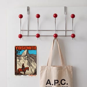 Op de afbeelding: Een verchroomde deurkapstok met acht haken met rode kogelvormige uiteinden. Onder de kapstok hangt een rechthoekige poster met een afbeelding van een persoon op een paard in Yosemite met de tekst YOSEMITE NATIONAL PARK California.