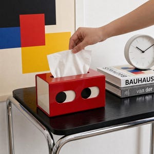 Puede incluir: Una caja para pañuelos rectangular con acabado en rojo brillante y dos aberturas circulares en el frente que revelan un interior blanco. La caja cuenta con una ranura superior de la cual se extrae un pañuelo blanco, diseño funcional.