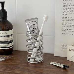 Peut inclure: Un porte-brosse à dents en métal argenté en forme de ressort cylindrique. À l'intérieur de la spirale, un tube de dentifrice Marvis argenté et une brosse à dents blanche sont placés verticalement. Le support est fait d'un fil continu.