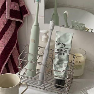 Pode incluir: Um organizador de casa de banho em fio cromado contendo uma escova de dentes elétrica verde sálvia com tampa transparente, uma escova de dentes manual cinzento-claro e um tubo de pasta de dentes Aesop. O objeto repousa sobre uma superfície branca.