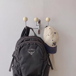 Può includere: Un appendiabiti da parete cromato con tre ganci a punta sferica color crema. Uno zaino Osprey grigio scuro è appeso al gancio sinistro, mentre un berretto da baseball color crema e blu navy con il logo dei New York Yankees pende dal gancio destro.