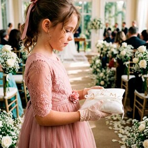 May include: A dusty rose lace and tulle dress with a matching floral waist accent, worn with beige lace gloves. The dress features elbow-length sleeves and a full skirt. A white rectangular ring pillow with the letters A & M is held in the hands.