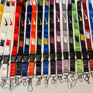Può includere: Una collezione di quindici cordini in tessuto di vari colori, tra cui giallo, bianco, rosso, nero, arancione, blu, verde, viola e grigio. Ogni cordino presenta il logo e la scritta Nike stampati ripetutamente, una fibbia in plastica nera e un gancio in metallo.