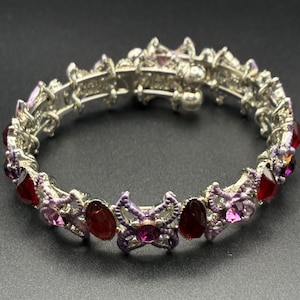May include: A silver-toned metal bracelet featuring a series of interconnected butterfly-shaped links. The design is accented with oval-shaped red gemstones and smaller round pink crystals, set within a delicate, textured, light purple metal frame.