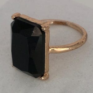 May include: A ring featuring a large, rectangular, faceted black stone set in a polished gold-tone metal band. The stone is held in place by four small prongs at the corners of the rectangular setting, creating a bold and geometric appearance.