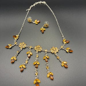 May include: A silver-toned necklace and matching earring set featuring amber-colored marquise and round crystals. The necklace has a delicate chain with floral-shaped clusters and dangling leaf-like crystal charms. The earrings are stud-style.