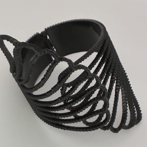 May include: A matte black cuff bracelet featuring an intricate, openwork design composed of multiple curved, textured wire-like segments that fan out from a central hinge, creating a layered, wing-like structure with a smooth inner band.