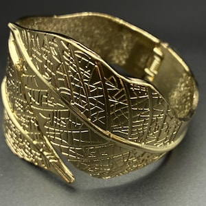 May include: A gold-toned metal cuff bracelet featuring a detailed, embossed leaf pattern across the entire outer surface. The design includes intricate, raised veins and a textured finish that mimics the natural structure of a large foliage leaf.